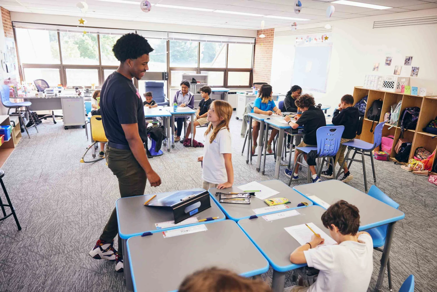 A teacher stands and talks to a student while other students sit at desks working in a brightly lit classroom.