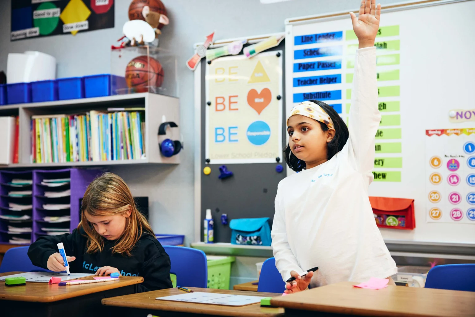 Two elementary school students sit at desks; one girl writes, and another raises her hand, in a colorful classroom with educational posters and supplies visible in the background.