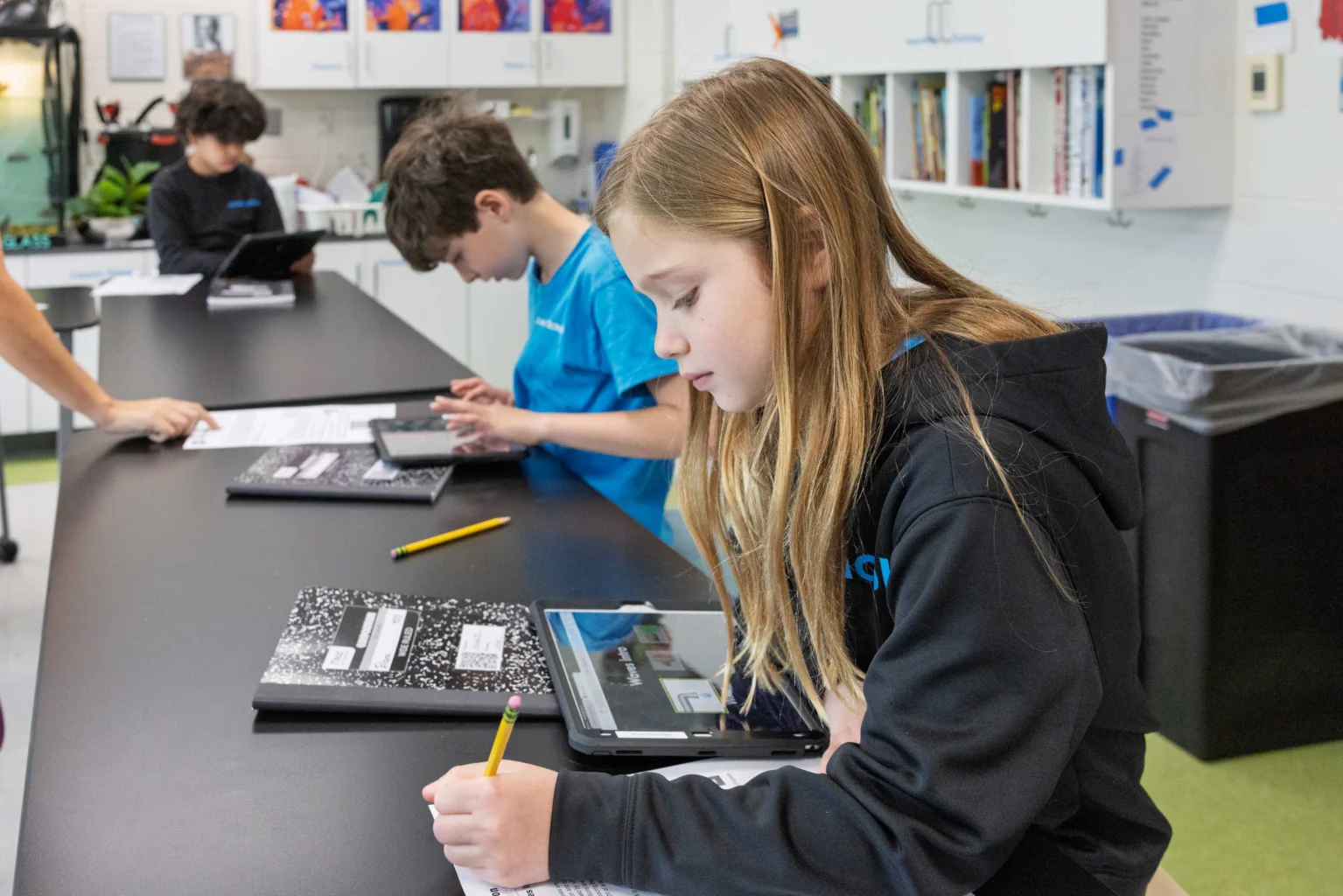 Three students sit at a classroom table, working on assignments with notebooks, pencils, and tablets in front of them. Shelves and a trash can are visible in the background.