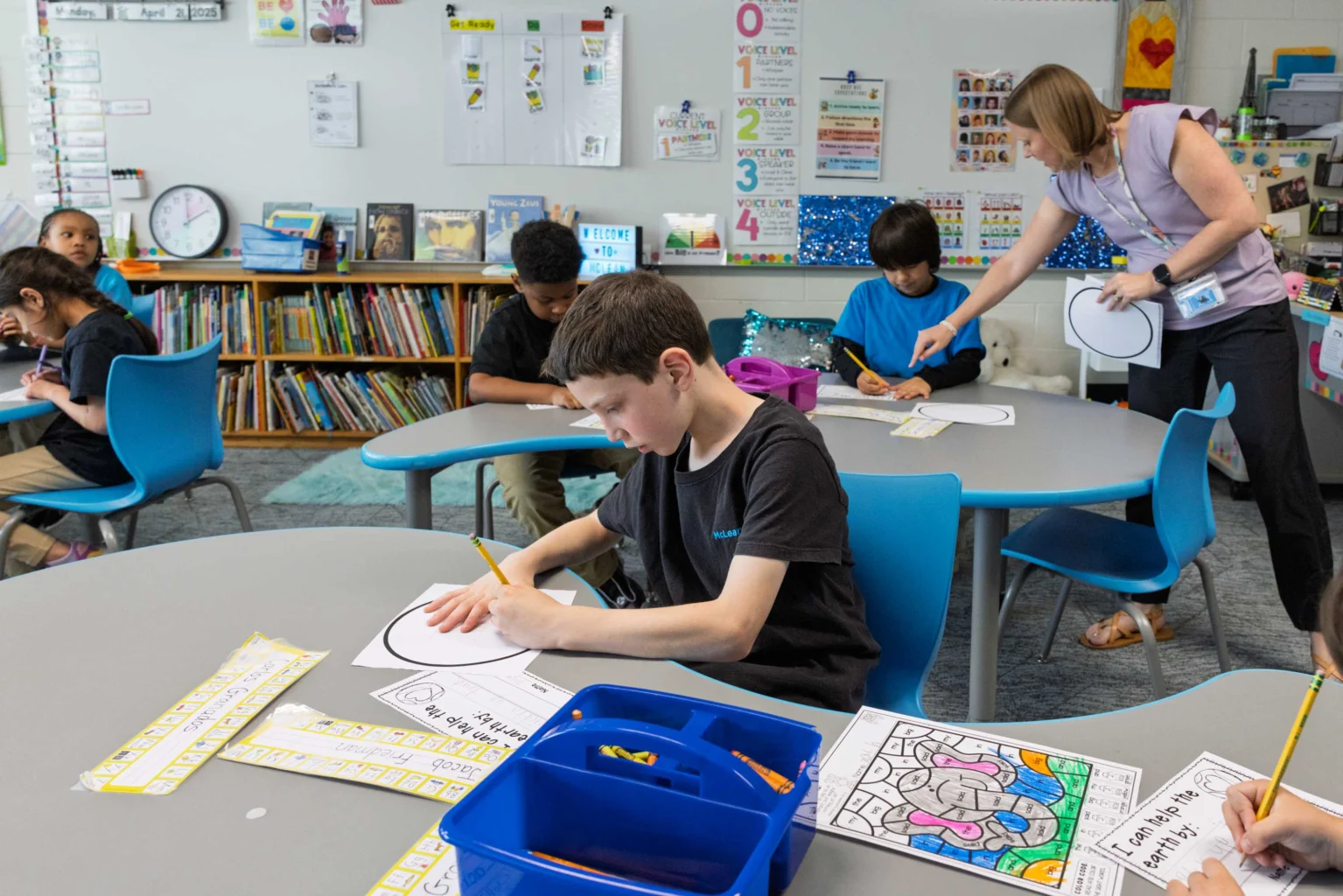Students sit at tables in a classroom working on coloring and writing activities while a teacher assists a student in the background.