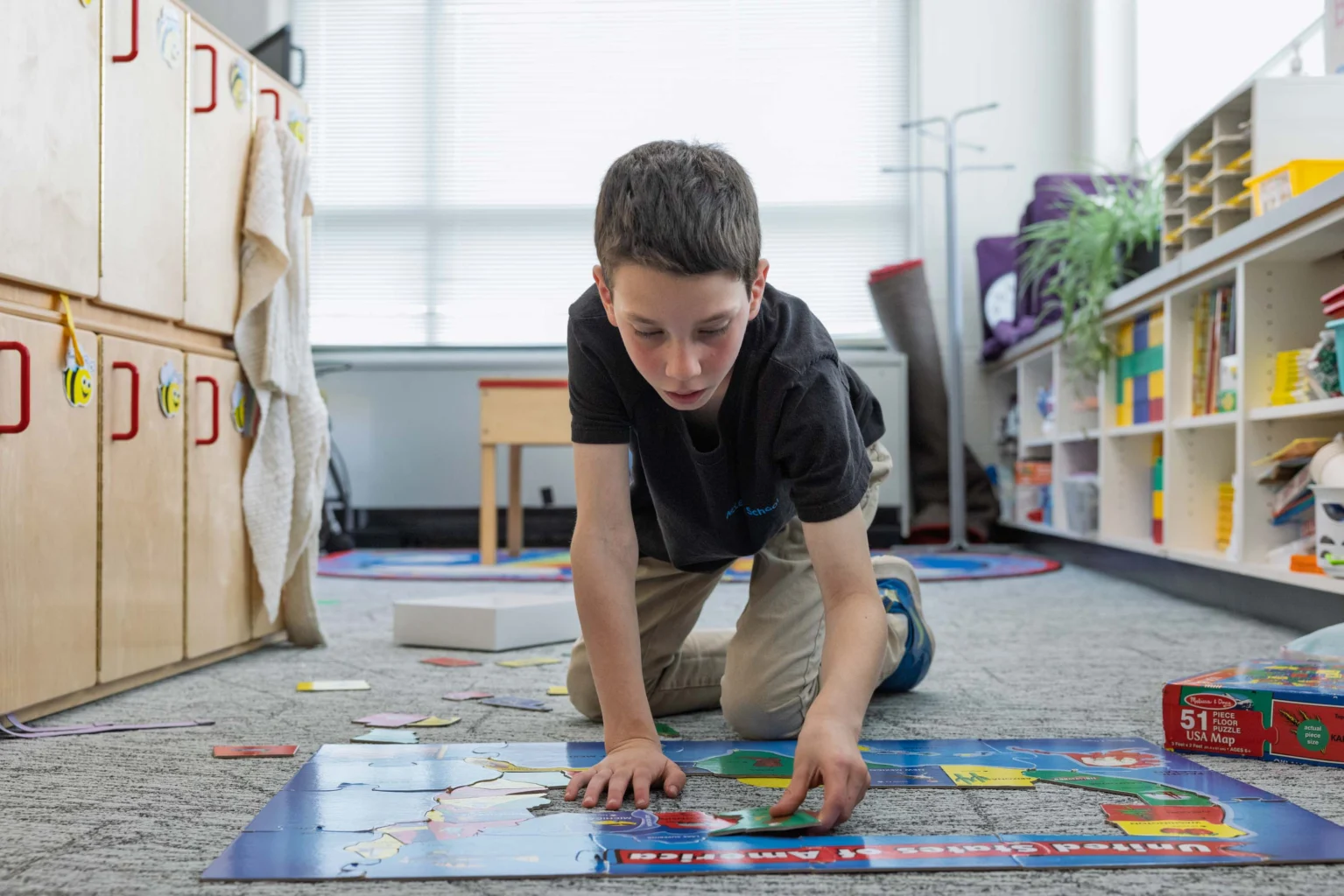 A boy kneels on the floor in a classroom, pointing at a large puzzle or map while other educational materials are scattered around him.