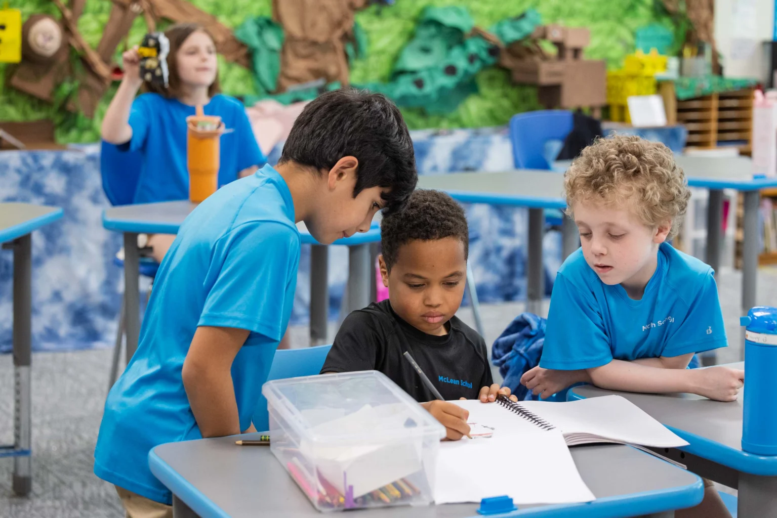Three children work together at a desk, looking at a sketchbook, while another child in the background holds an object in a classroom setting.