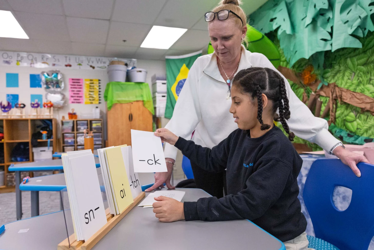A teacher assists a student with phonics flashcards at a classroom table, focusing on word endings like "--ck" and "at--".