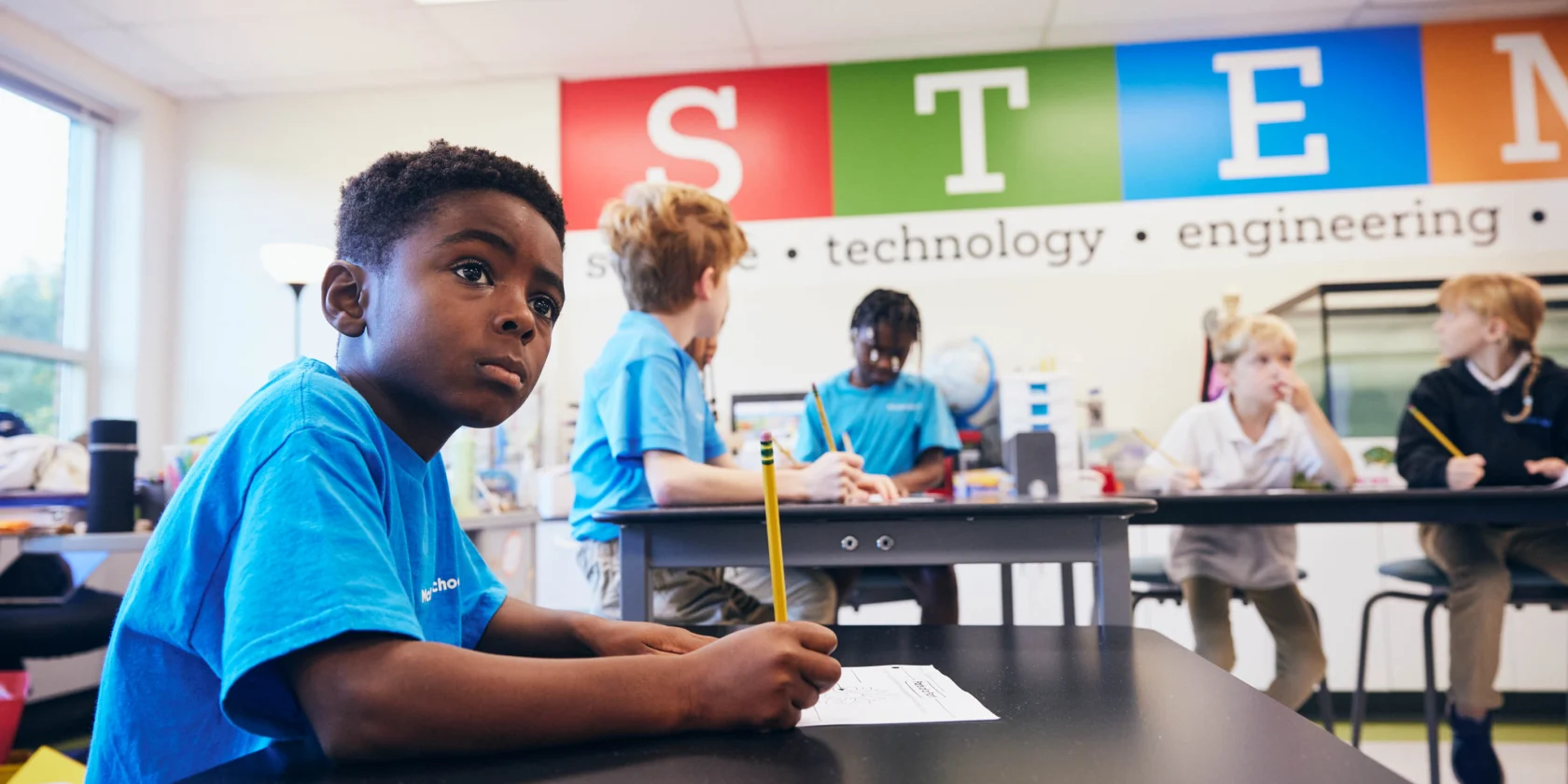 A boy sits at a desk holding a pencil in a classroom with STEM posters on the wall, while other students interact in the background.