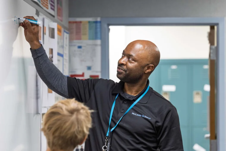 A teacher writes on a whiteboard while a student stands nearby in a classroom with lockers and colorful posters on the walls.