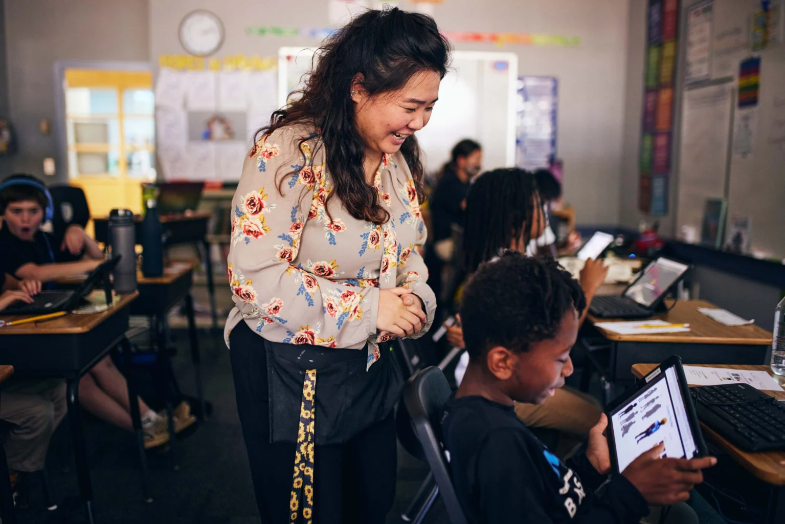 A teacher smiles and stands beside a student using a tablet in a classroom filled with students working on laptops and tablets.