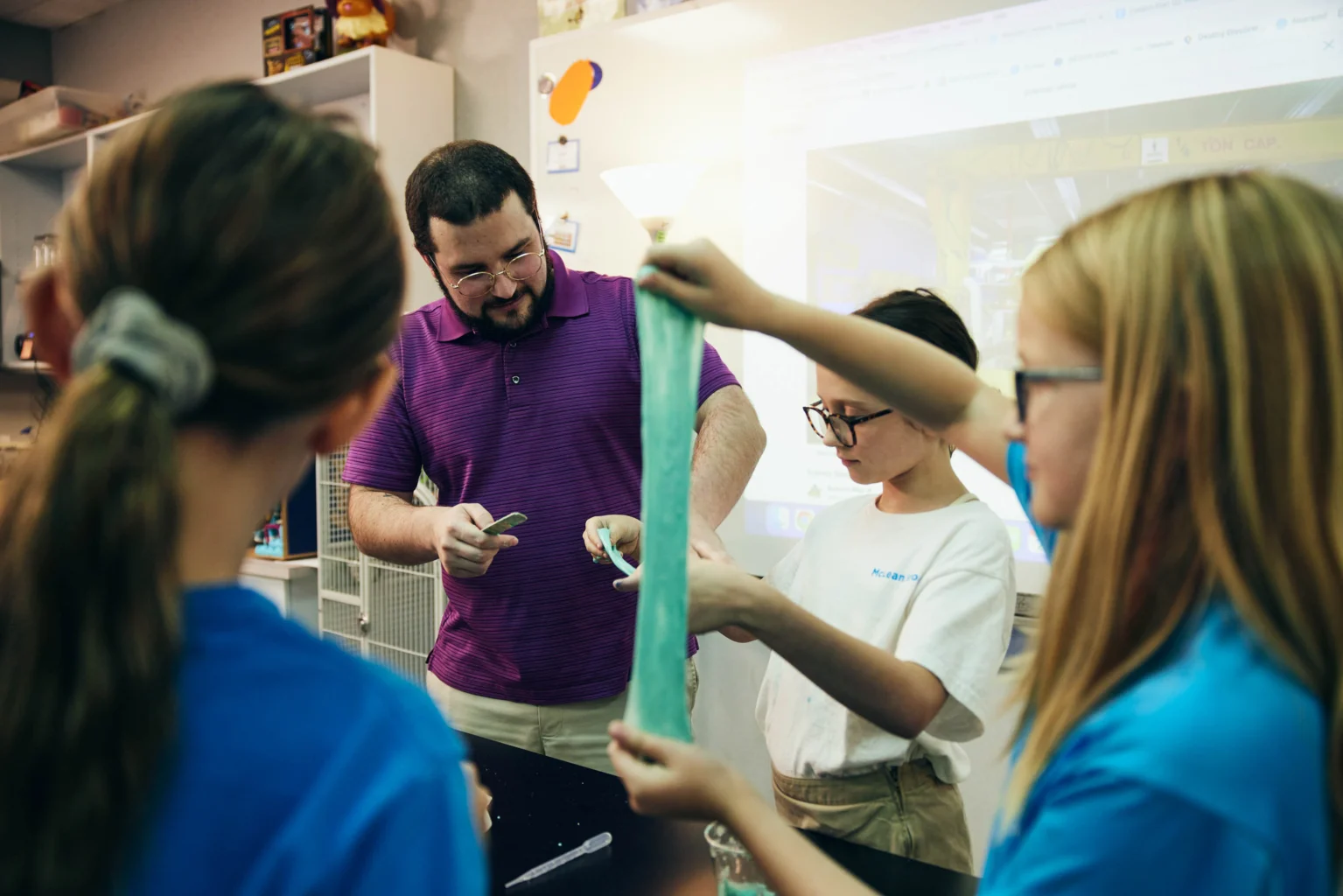 A teacher in a purple shirt stands with three students in a classroom as they stretch and examine a piece of green slime.