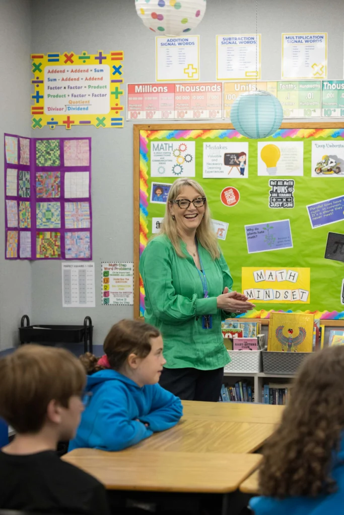 A teacher stands and smiles at the front of a classroom, with students seated at desks and colorful educational posters on the walls.