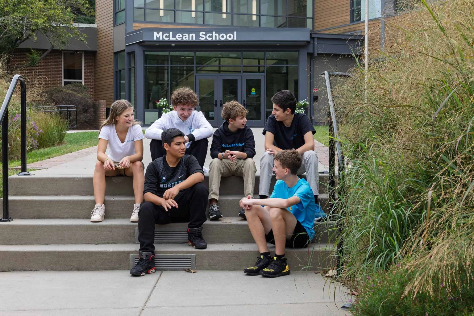 Five teenagers sit and talk on the steps outside McLean School, with the school building and entrance visible in the background.
