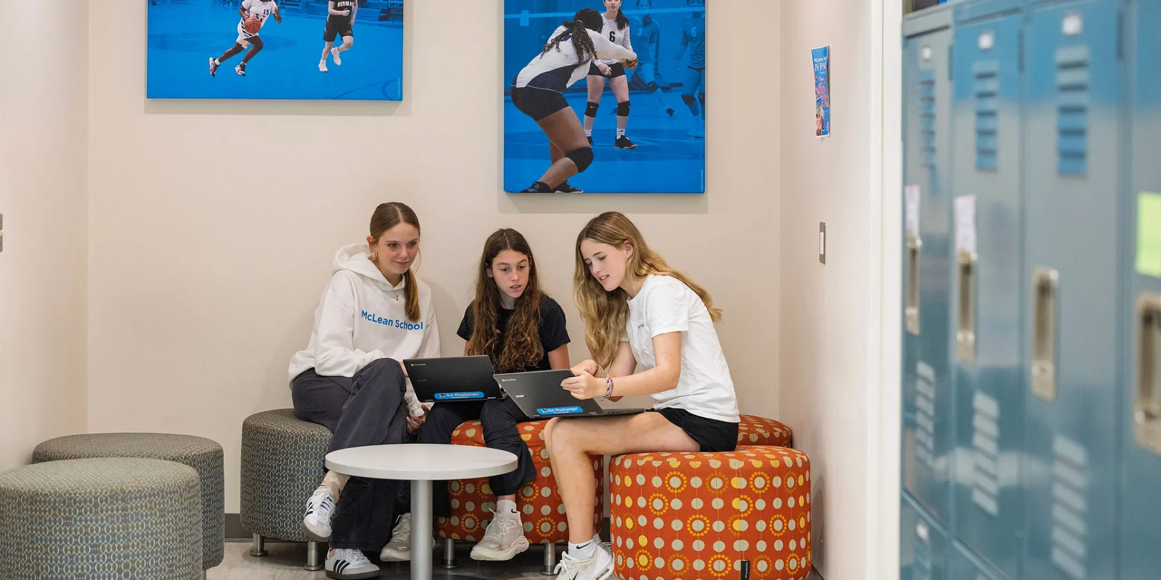 Three young women sit around a small table with laptops in a school hallway, near lockers and sports-themed wall art.