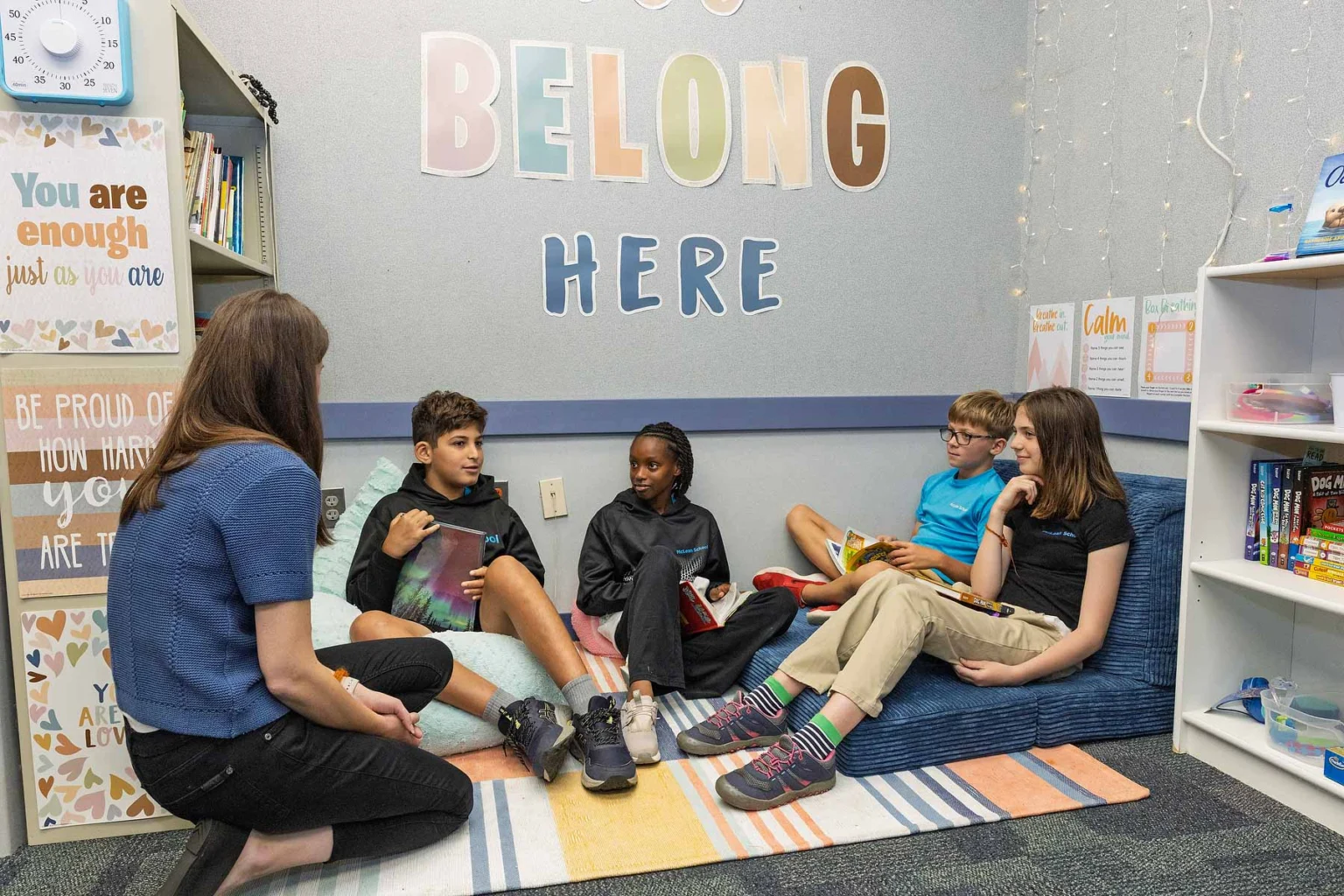 Five people, including a teacher and four students, sit on cushions in a classroom reading corner under a "You Belong Here" sign, surrounded by books and motivational posters.
