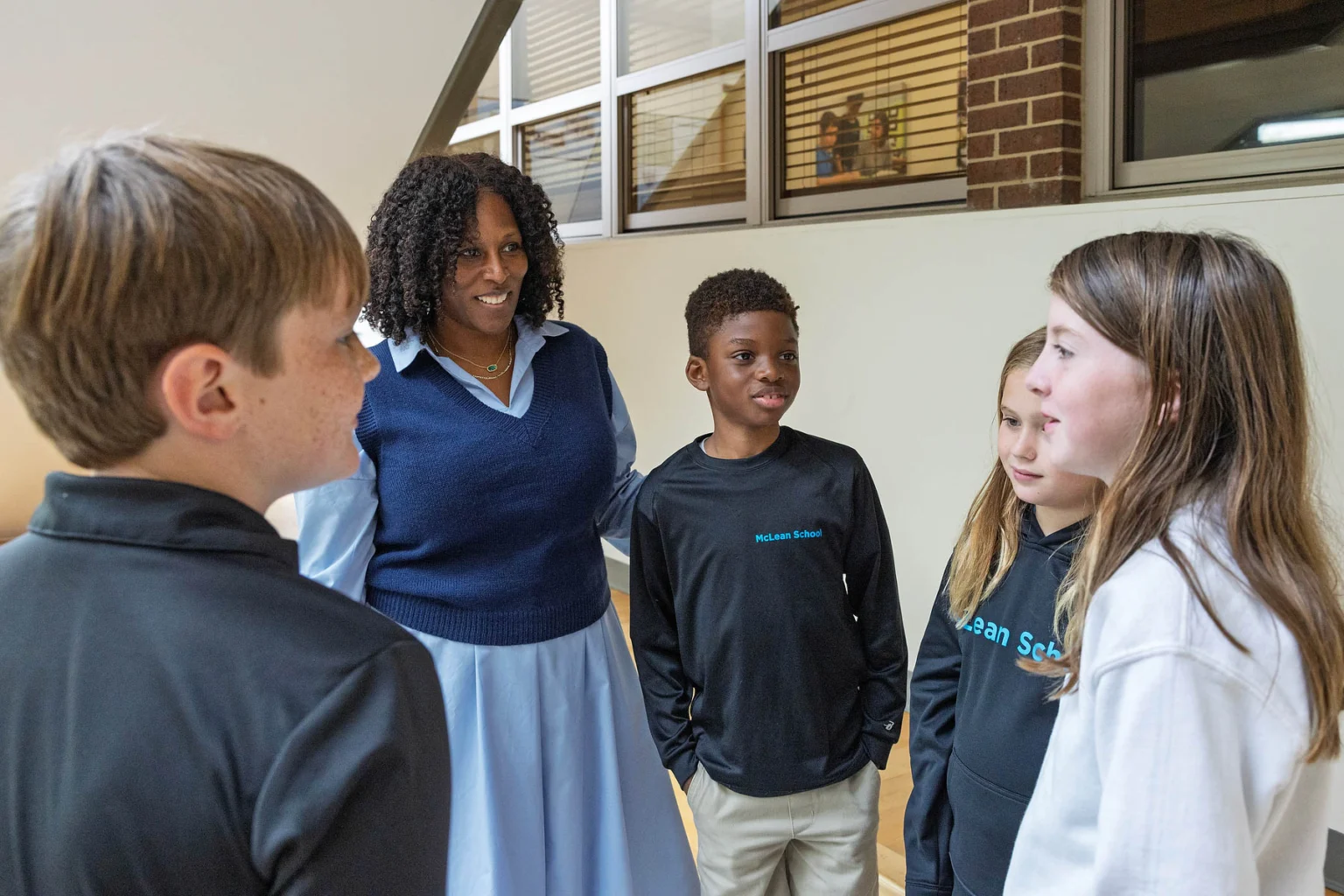 A teacher stands with four students in a hallway, engaging in conversation. The group appears to be in a school setting.