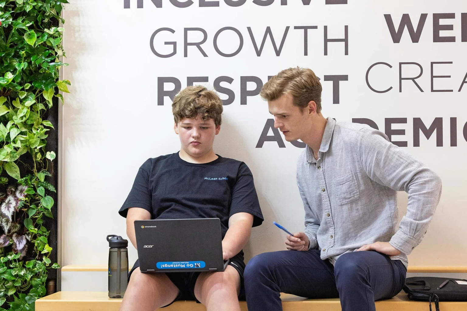 A student and an adult sit on a bench looking at a laptop together; motivational words are displayed on the wall behind them.