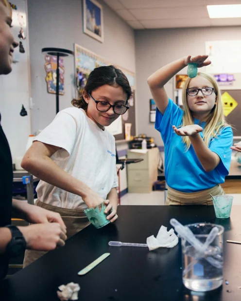 Four students at a classroom table perform a science experiment with green and blue slime, using cups and mixing tools. Other students and classroom materials are visible in the background.