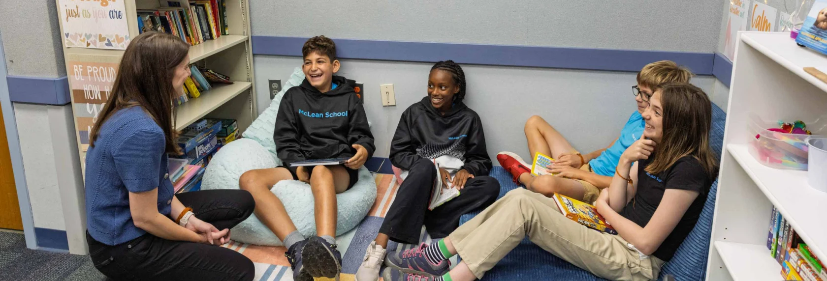 A teacher sits on the floor with four students in a classroom reading corner. The wall behind them says "You Belong Here." The group appears engaged and relaxed.
