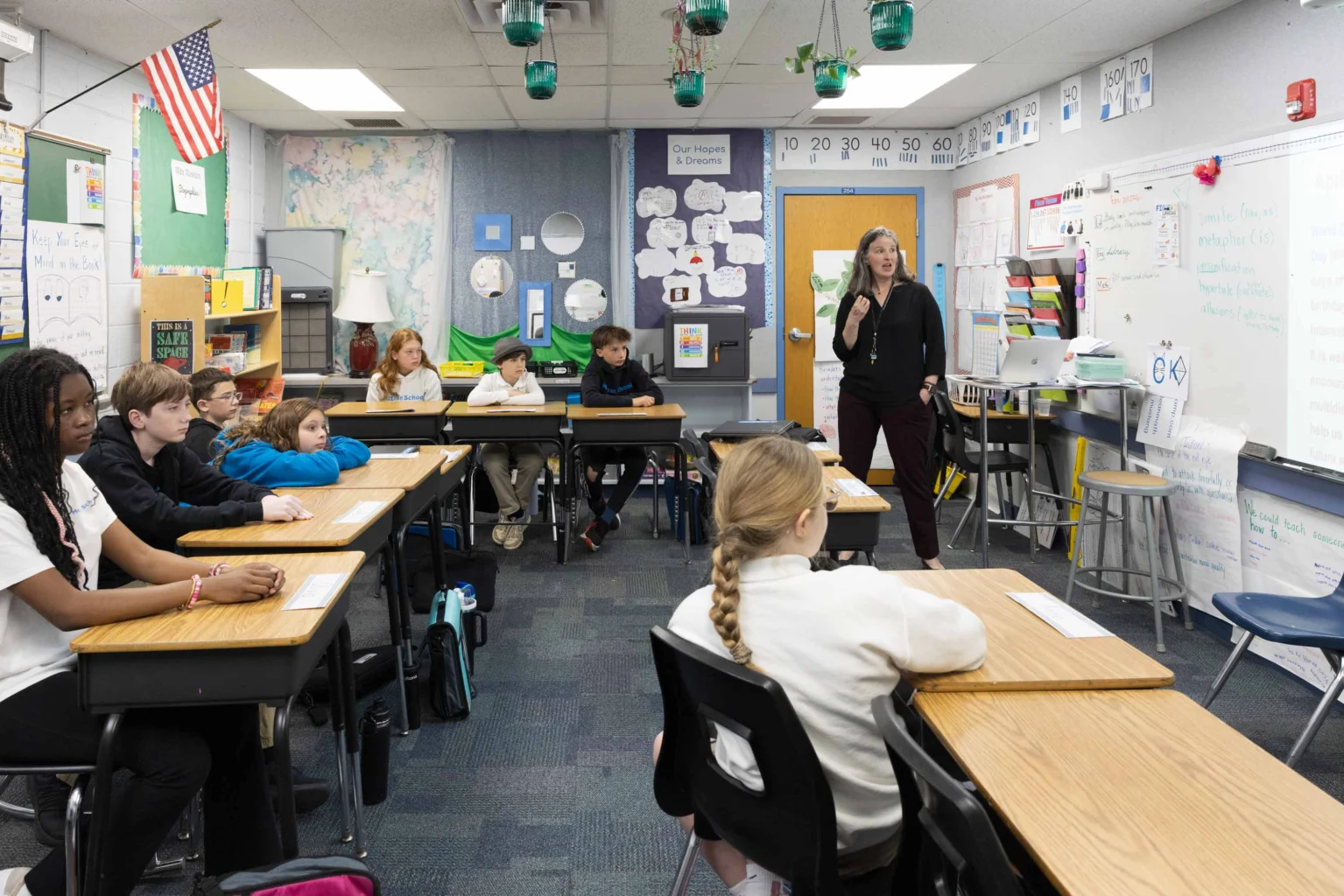 A teacher stands at the front of a classroom speaking to students seated at desks arranged in a U-shape. Educational materials and posters decorate the walls.