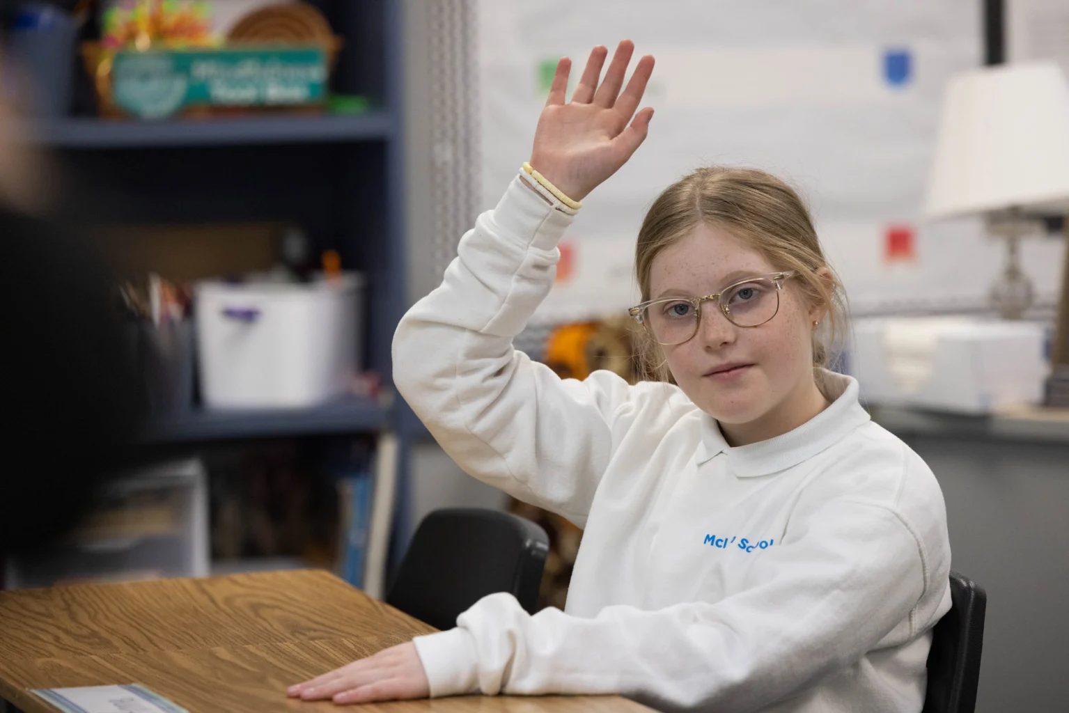 A student with glasses and a white sweatshirt raises her hand while sitting at a desk in a classroom.