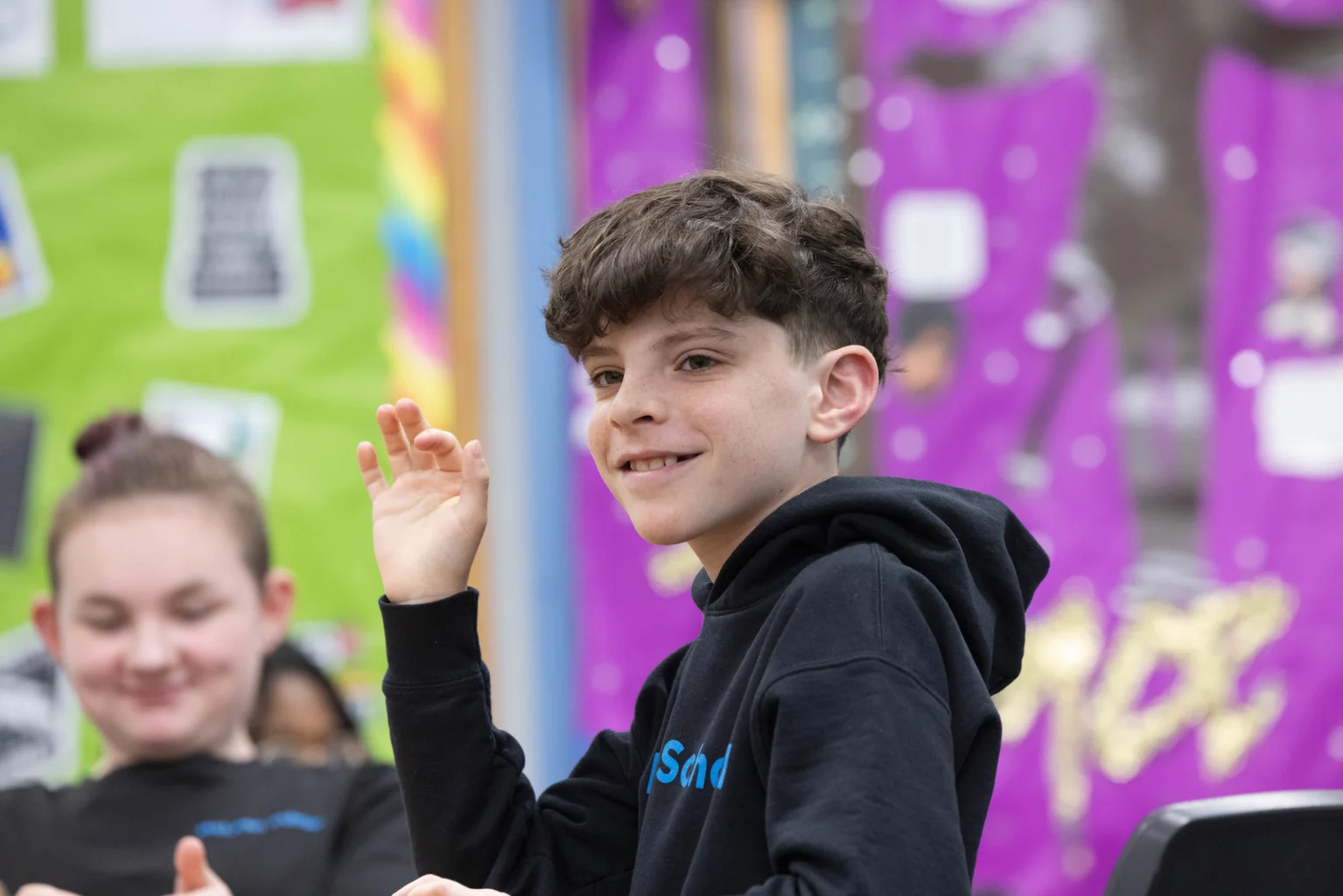 A boy with short brown hair wearing a black hoodie raises his hand while sitting in a classroom. Another student is blurred in the background.