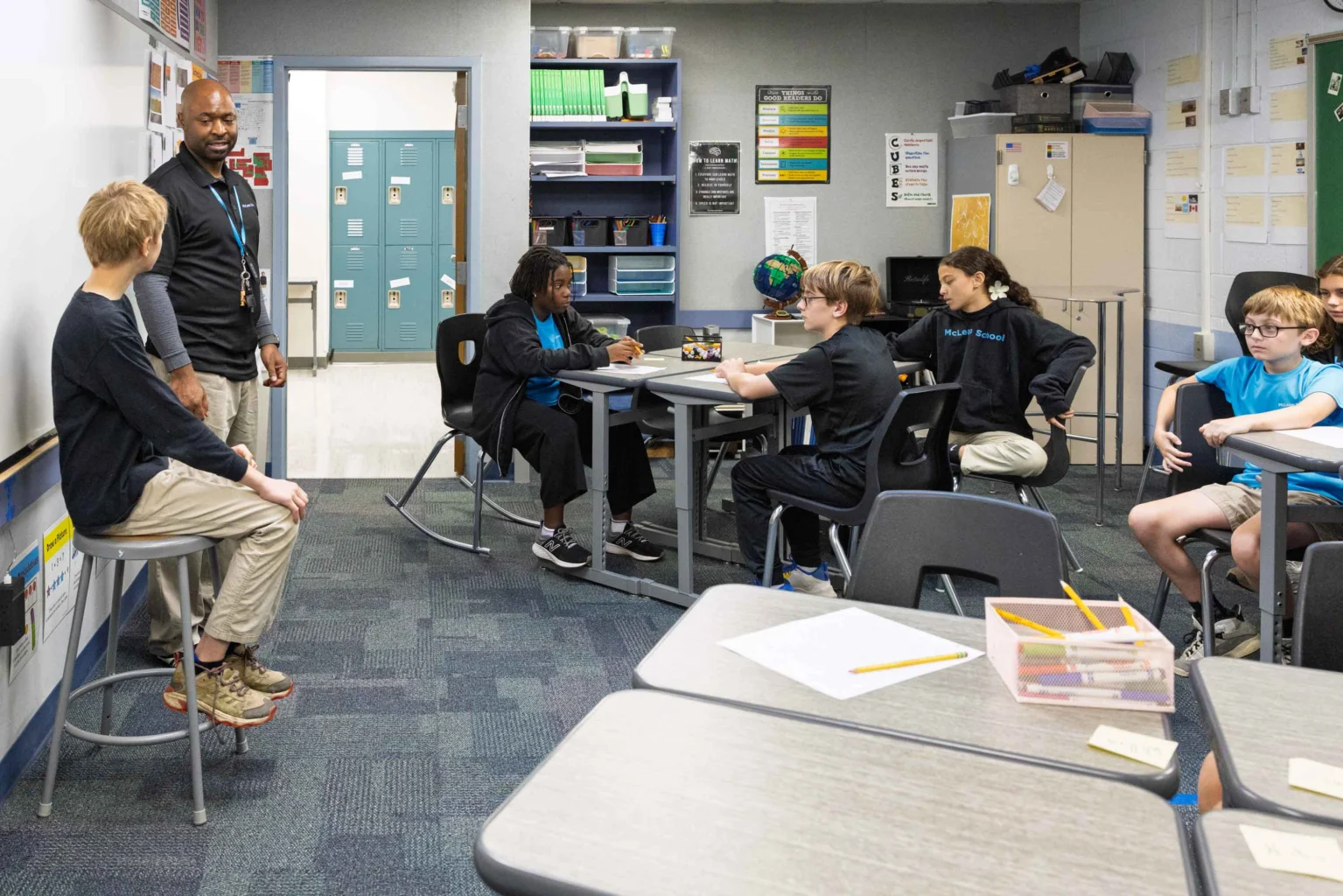 A teacher stands at the front of a classroom while students sit at desks in groups, listening and participating in a discussion.