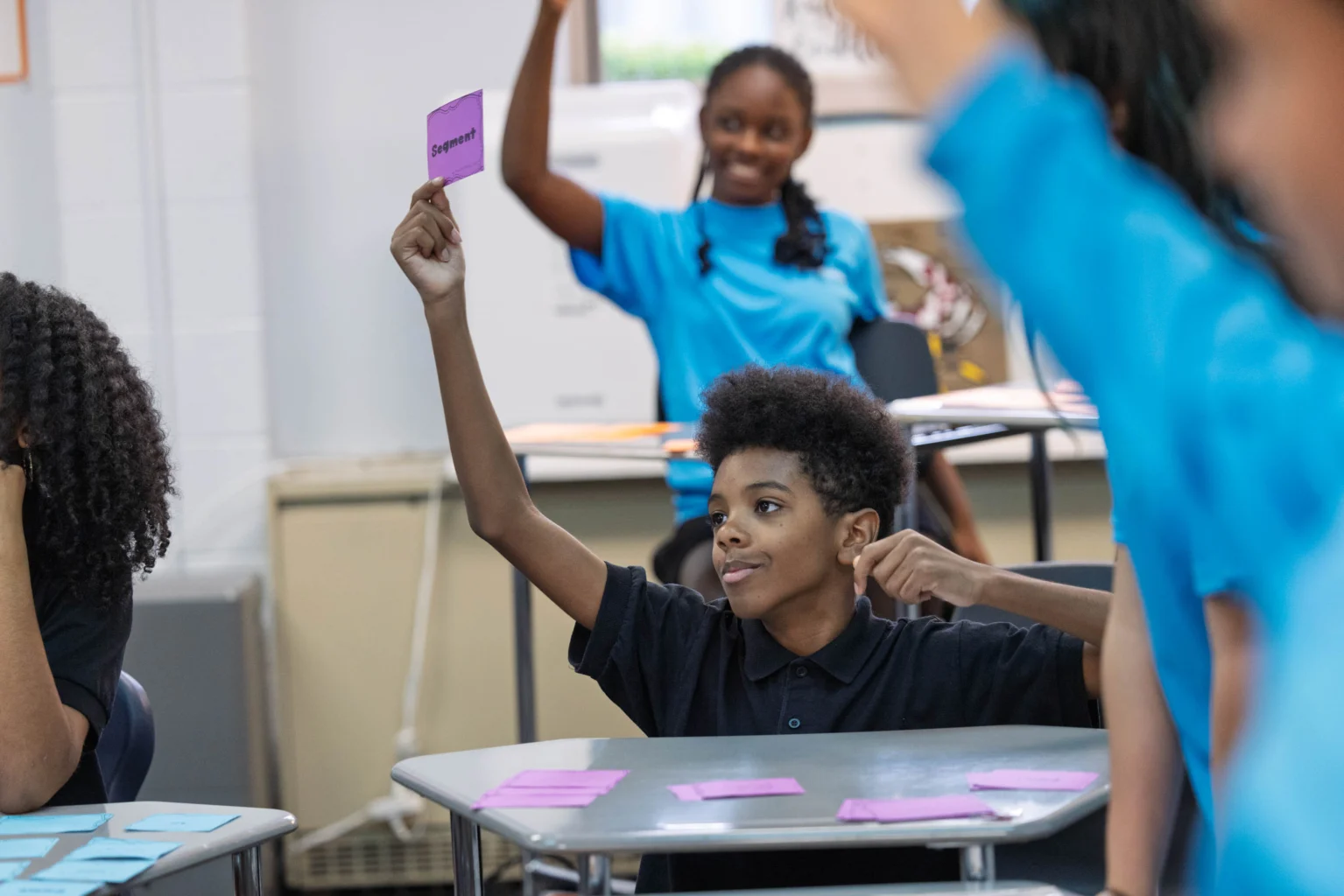 A student seated at a desk raises a purple card, while another student in the background also raises their hand in a classroom setting.