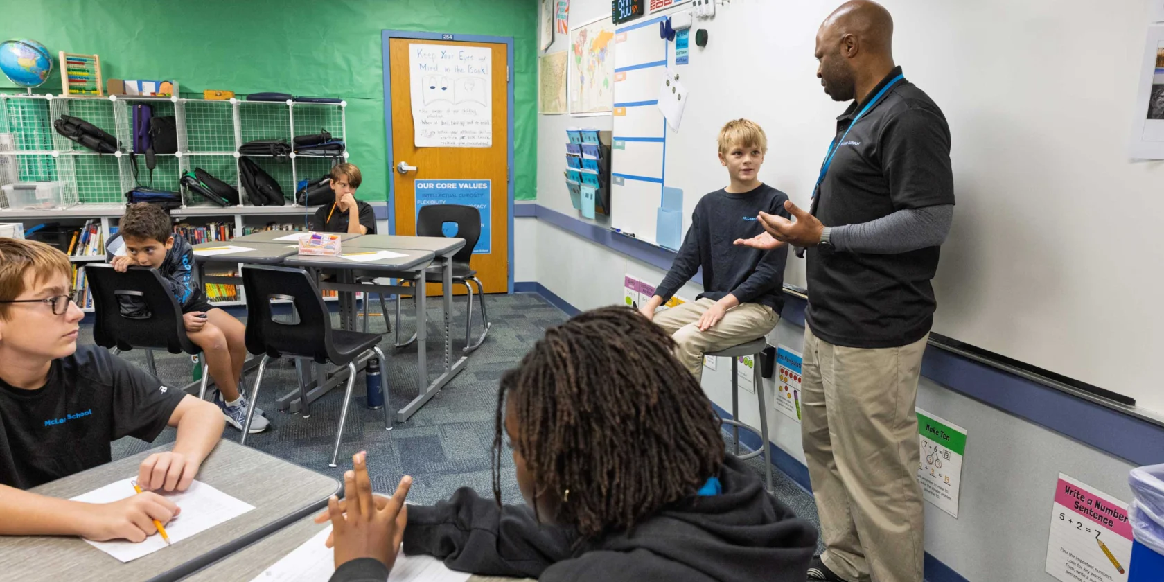 A teacher stands at the front of a classroom speaking to students, with one student sitting on a table near the whiteboard. Other students sit at desks with papers and pencils.