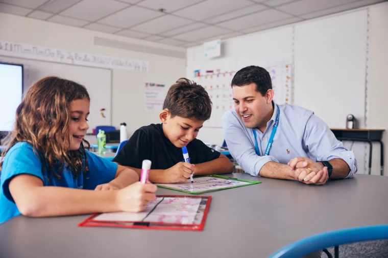 A teacher sits at a table with two students who are writing on whiteboards in a classroom.