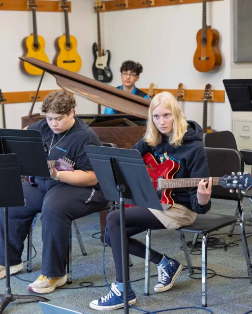 Three students play guitars and piano in a music classroom, seated with music stands in front of them; guitars hang on the wall in the background.