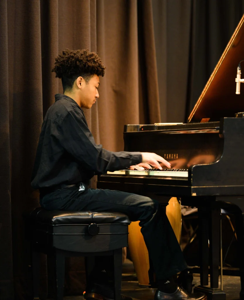 A young person in black clothing plays a Yamaha grand piano on stage, seated on a piano bench with a dark curtain in the background.