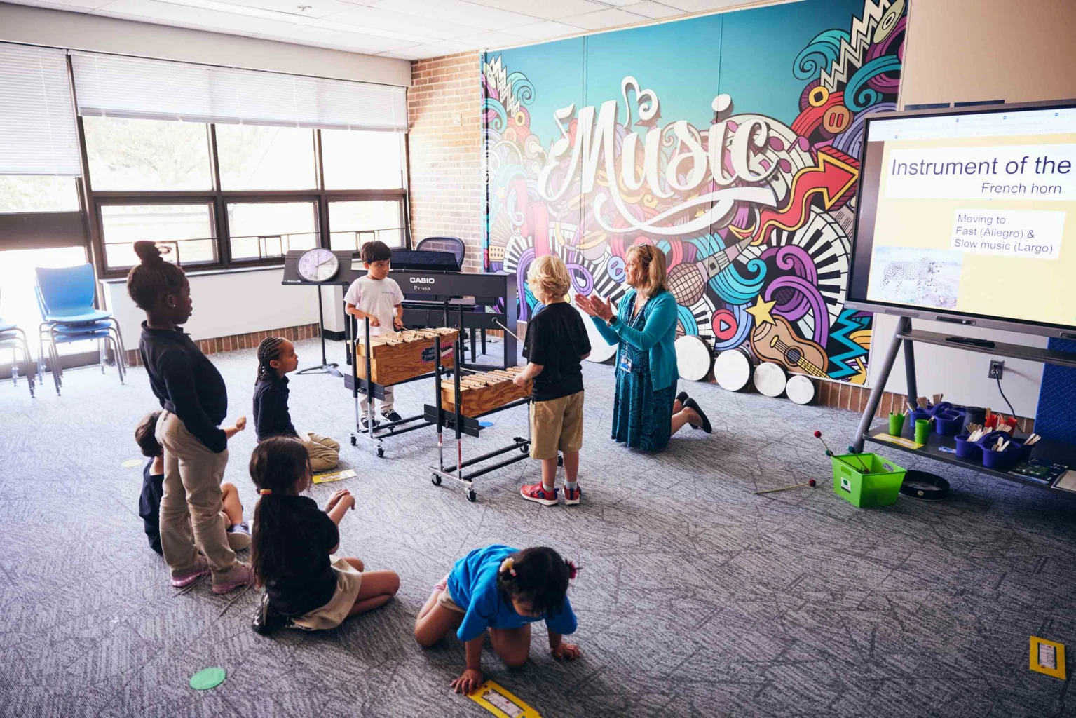 A group of young children participate in a music class, some sitting and some standing, while a teacher instructs near a large “Music” mural and a digital whiteboard.