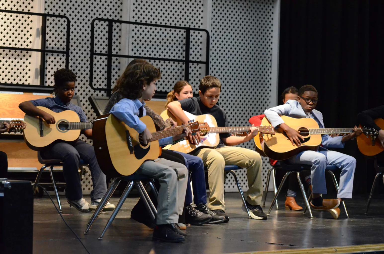 A group of children sit in chairs on stage, each playing acoustic guitars during a performance or rehearsal.