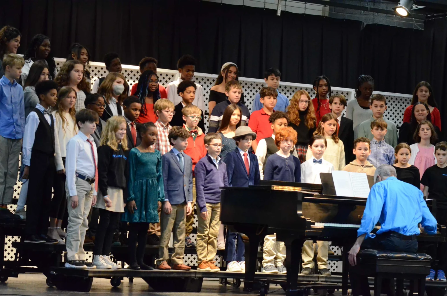 A children's choir stands on risers and sings while a person in a blue shirt plays the piano, facing them, on a stage with a black curtain backdrop.
