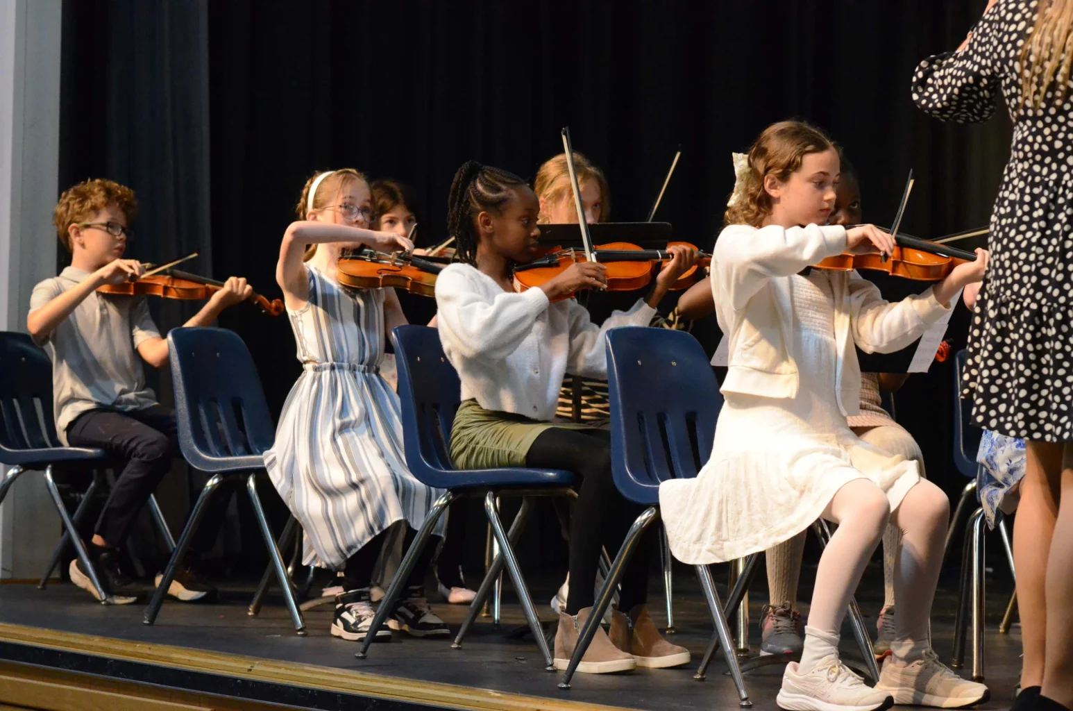 A group of children sit on stage playing violins, following the direction of an adult standing in front of them.