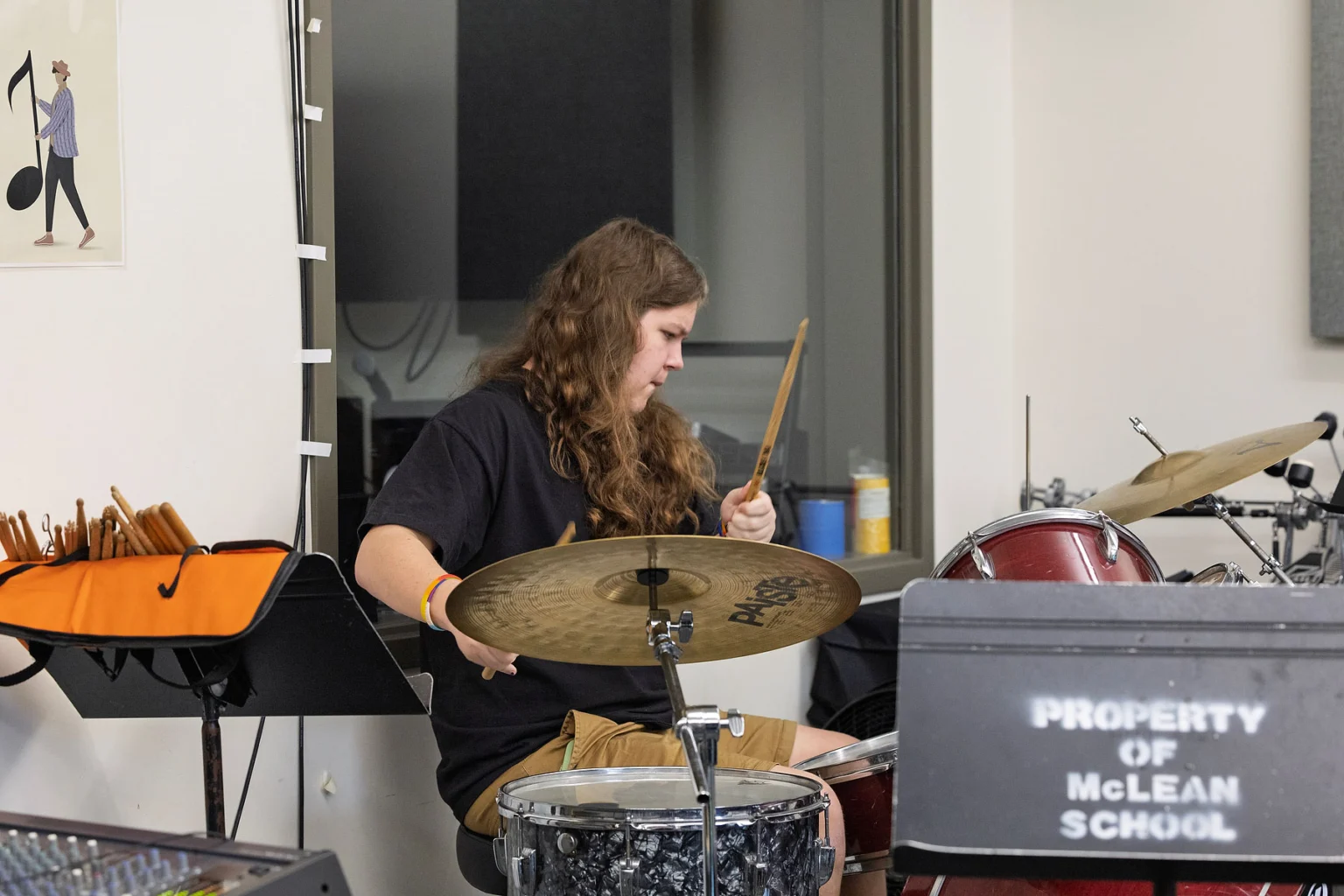 A person plays a drum set in a music room with a sign that reads "Property of McLean School" visible in the foreground.