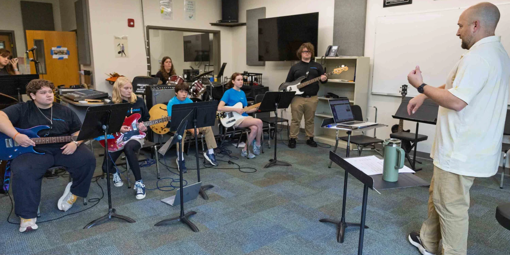 A music teacher stands at the front of a classroom instructing a group of students playing electric guitars and bass guitars. Music stands are arranged in front of the seated students.