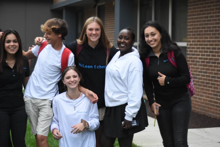 Five students pose and smile outdoors near a brick building, wearing casual clothes and backpacks.