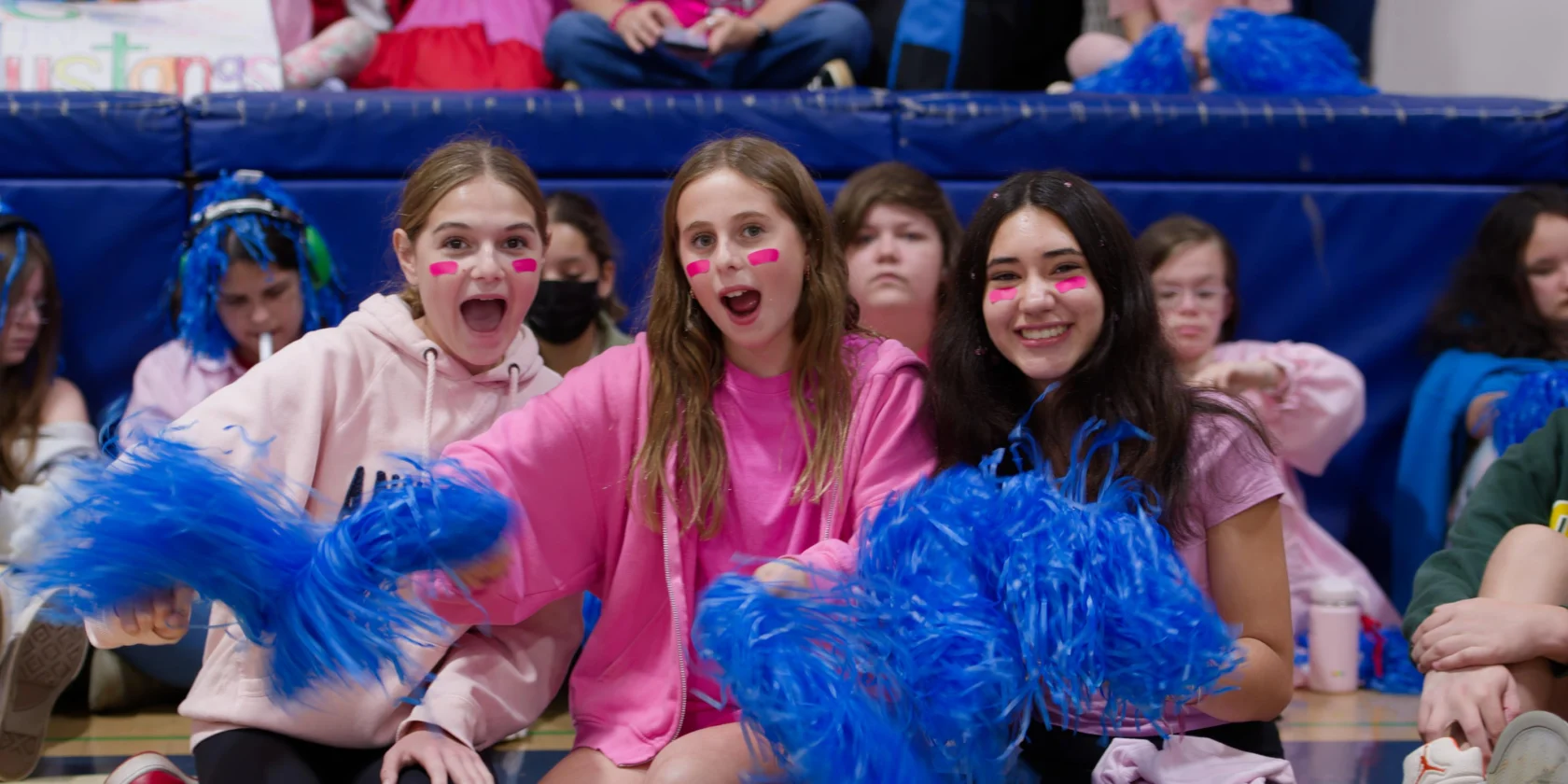 Three girls wearing pink shirts and face paint sit on a gym floor holding blue pom-poms, surrounded by other students in the background.