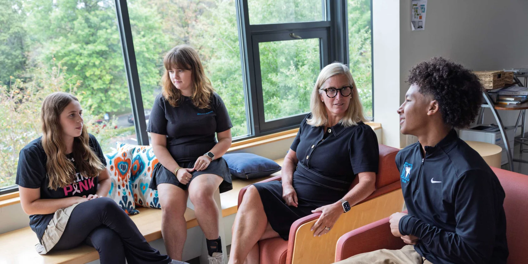 Four people sit and talk in a brightly lit room with large windows, two on a bench and two on chairs, appearing engaged in conversation.