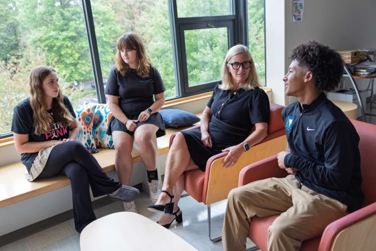 Four people sit and talk in a brightly lit room with large windows, two on a bench and two on chairs, appearing engaged in conversation.