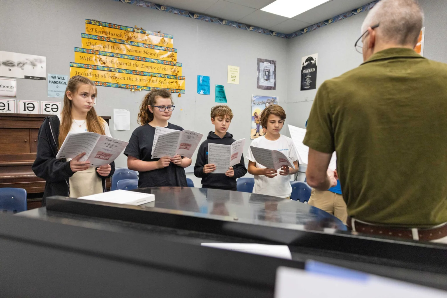 A group of students stand holding music sheets and sing while a teacher, seen from behind, leads them in a classroom.