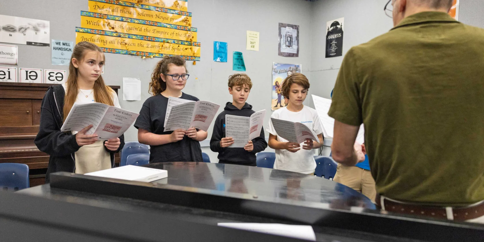 A group of students stand holding music sheets and sing while a teacher, seen from behind, leads them in a classroom.