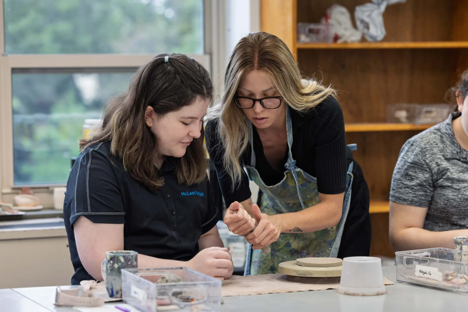 An instructor helps a student shape clay on a pottery wheel in a classroom, with shelves and supplies in the background.