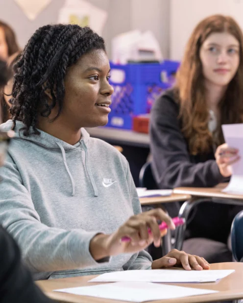 A student in a gray hoodie speaks while holding a pen in a classroom, with other students sitting at desks and listening.