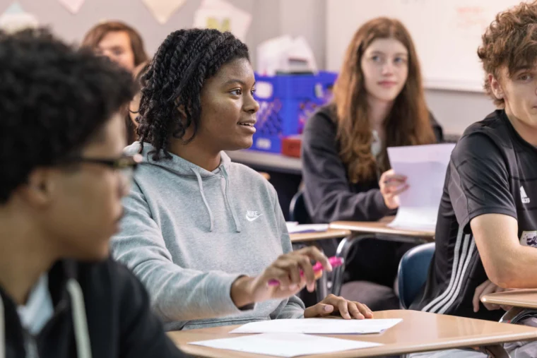 A student in a gray hoodie speaks while holding a pen in a classroom, with other students sitting at desks and listening.