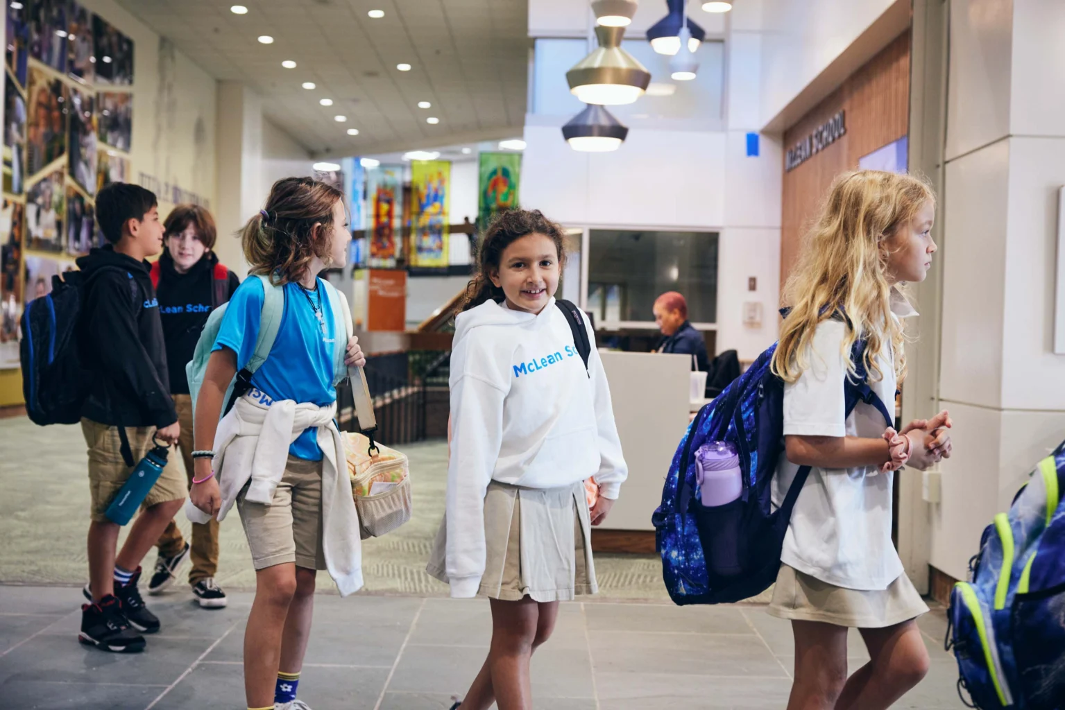 A group of elementary school students in uniforms walk through a modern school hallway with backpacks and water bottles.