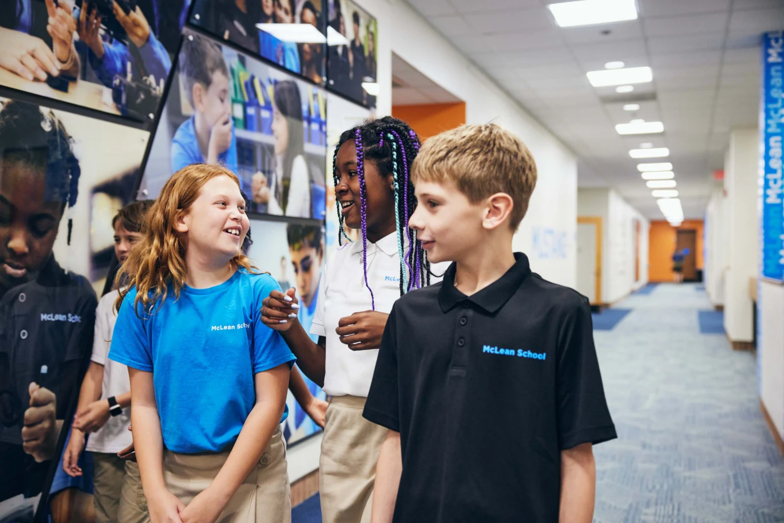 Four elementary-aged students in school uniforms walk and talk together in a hallway decorated with educational photos and blue accents.