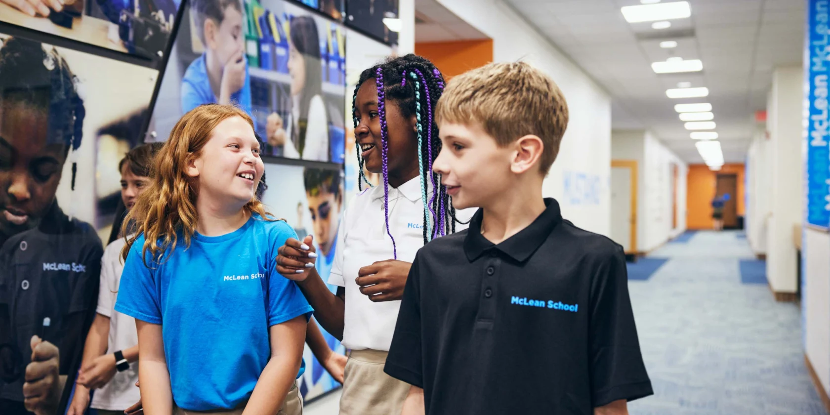 Four elementary-aged students in school uniforms walk and talk together in a hallway decorated with educational photos and blue accents.