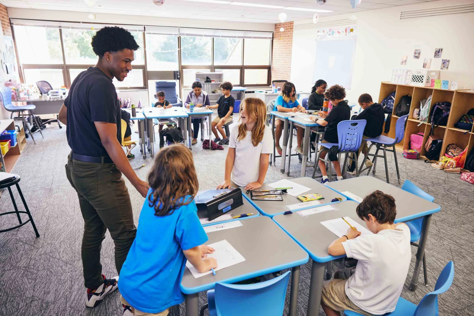 A teacher speaks with students working at tables in a bright classroom, while other students are seated at desks in the background.