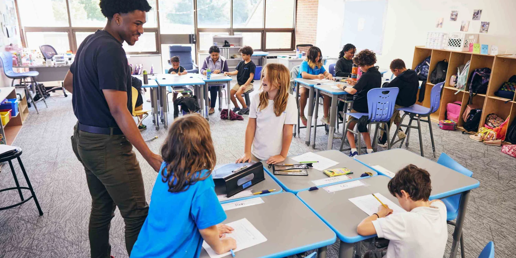 A teacher speaks with students working at tables in a bright classroom, while other students are seated at desks in the background.