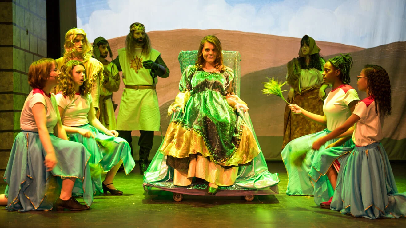 A woman in a green medieval dress sits on a throne surrounded by people in costumes, some kneeling, on a stage set with a castle and landscape backdrop.