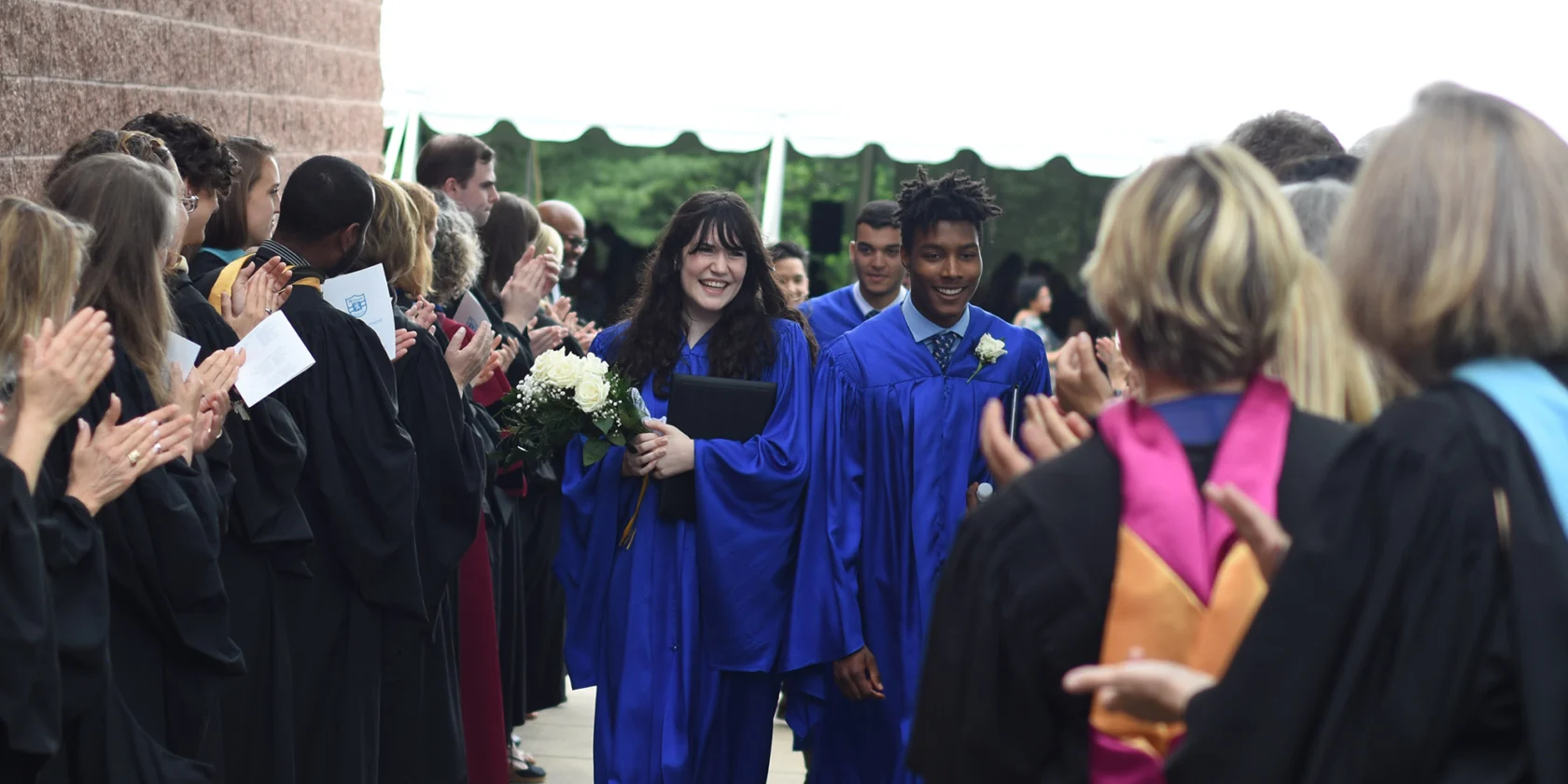 Two students in blue graduation gowns walk down an outdoor aisle while faculty and others in academic regalia applaud on either side.